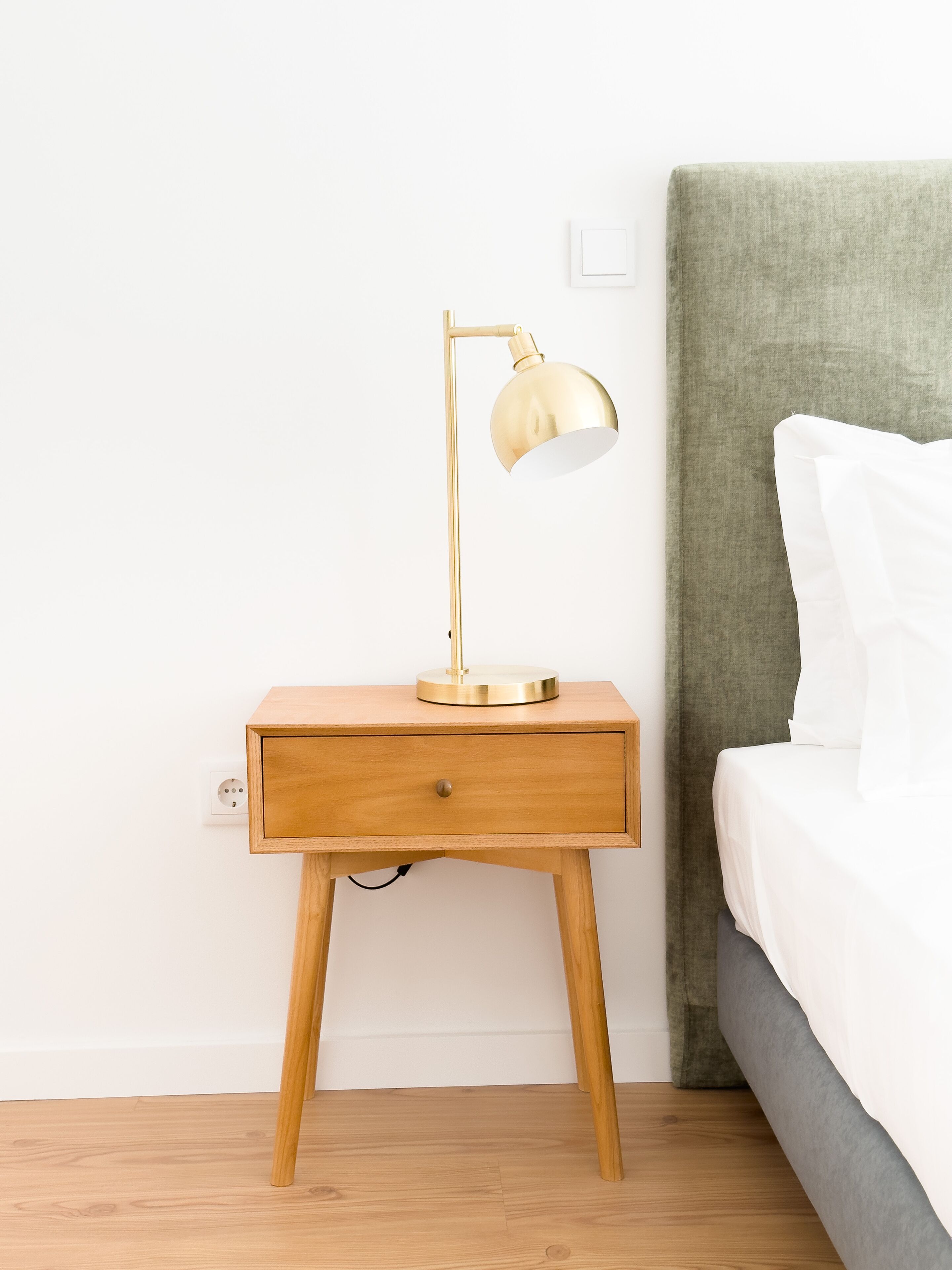 Een houten nachtkastje met een gouden lamp, een lade en een stopcontact.