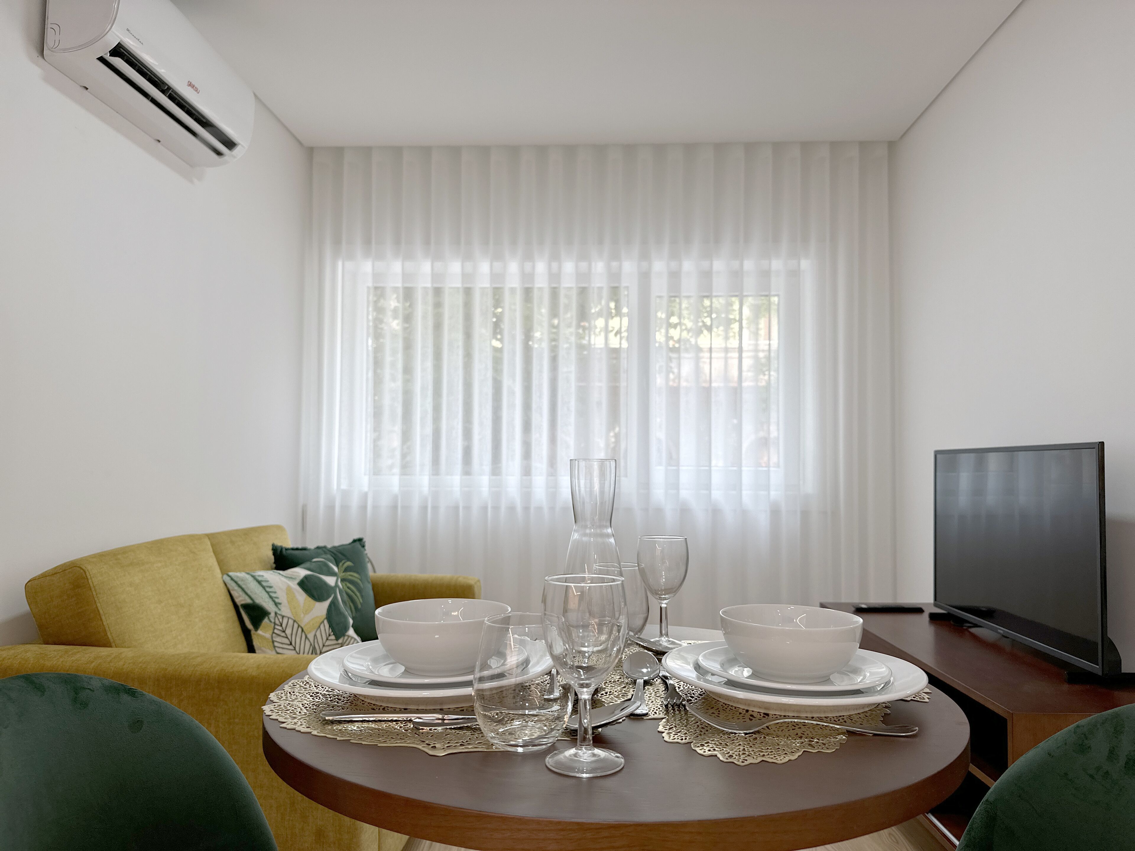 A dining area with a round table set for four, a sofa, a TV, and a window with sheer curtains.