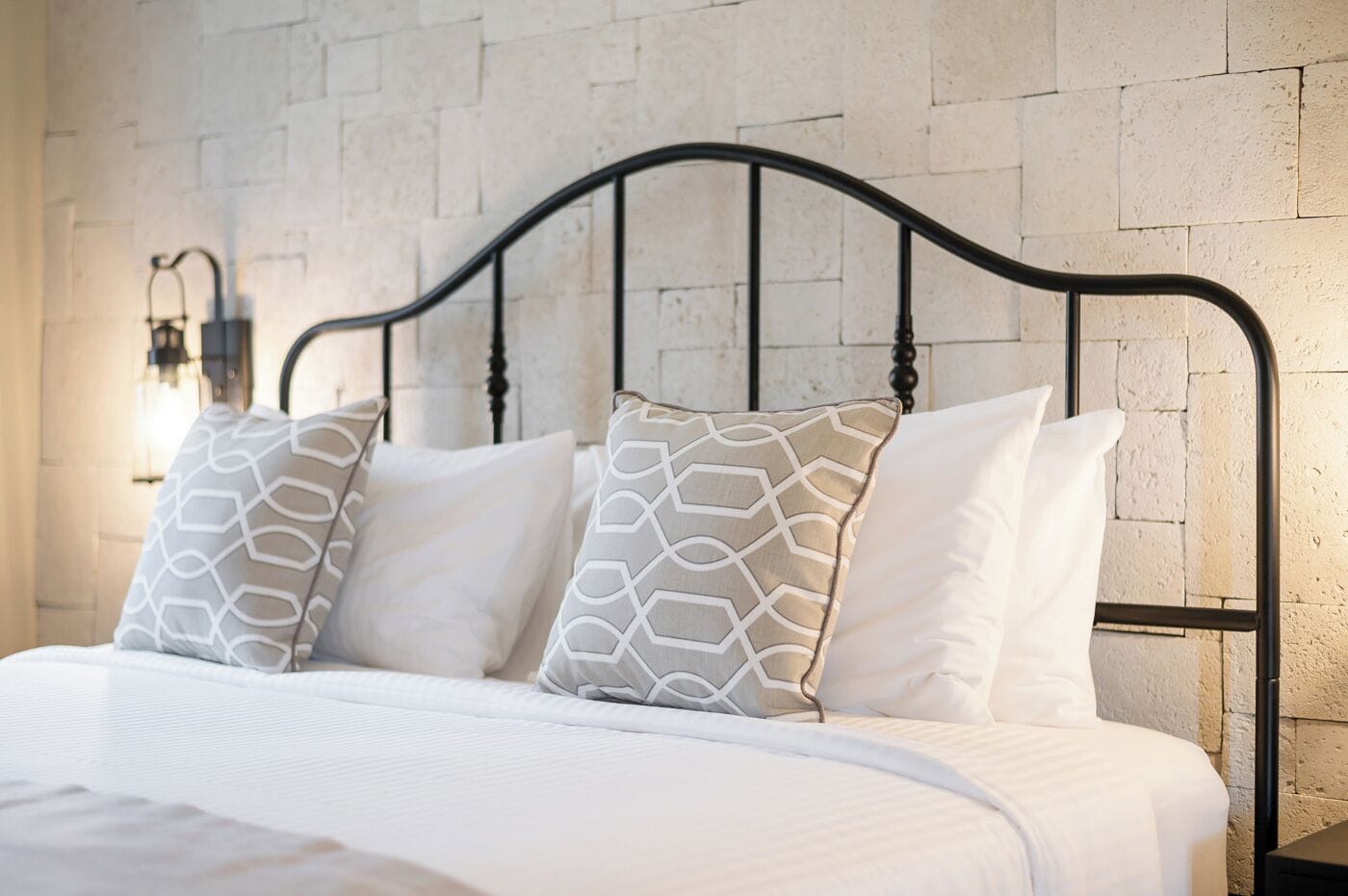 Een bed met een witte matras en twee gestreepte kussens, een zwart metalen hoofdbord en een muur met een reliëf stenen patroon.