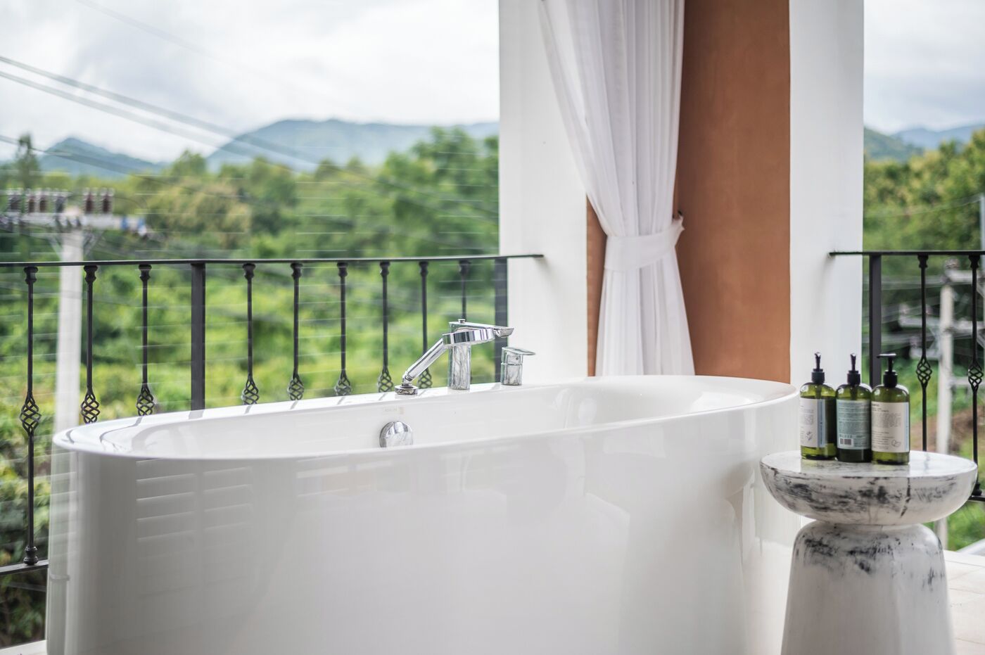 A large, white freestanding bathtub with a modern faucet, positioned on a balcony with a metal railing. In the background, there is a view of lush green mountains.