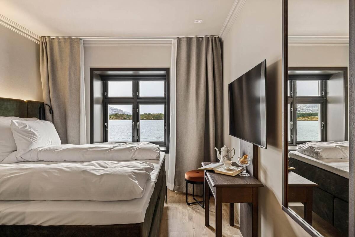 Habitación de hotel con dos camas, un escritorio con silla, televisor y ventanas amplias con vista al lago.