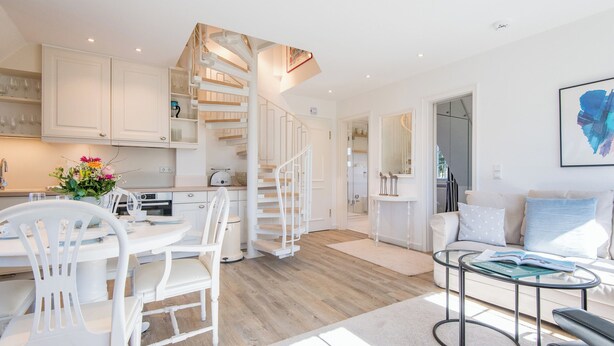 Eine moderne Küche mit weißen Schränken, ein Essbereich mit weißen Stühlen, eine Wendeltreppe und ein Wohnzimmer mit einem Glastisch.