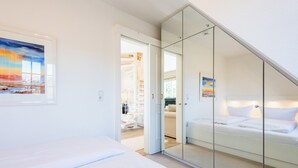 Ein modernes Schlafzimmer mit einem großen Bett, einer Glastrennwand und Blick auf eine Treppe, die in einen Wohnbereich führt.