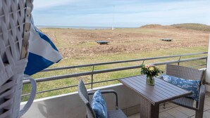 A balcony with wicker furniture, a table with a vase, and a view of a field with a wind turbine. - 1-zimmerwohnung mit Meerblick und ca. 46 m² Wohnfläche, für bis zu 2 Personen (Wenningstedt-Braderup (Sylt))