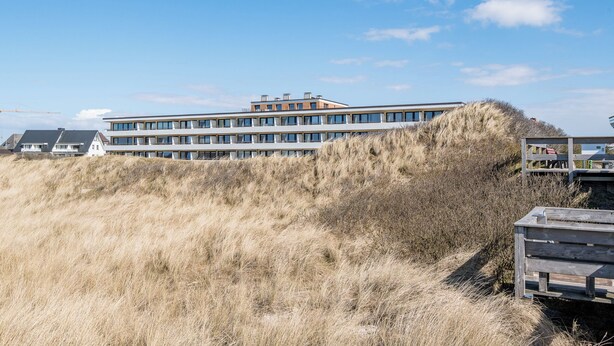 A modern multi-story building with a flat roof, situated on a grassy hill with a clear sky. - 1-zimmerwohnung mit Meerblick und ca. 46 m² Wohnfläche, für bis zu 2 Personen (Wenningstedt-Braderup (Sylt))