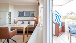 A modern living room with a sofa, dining table, and chairs, a balcony with a view, and a small kitchen area. - 1-zimmerwohnung mit Meerblick und ca. 46 m² Wohnfläche, für bis zu 2 Personen (Wenningstedt-Braderup (Sylt))