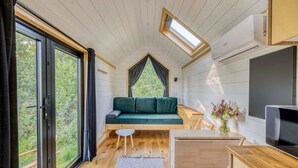 Interior acogedor de una cabaña con cama en el altillo, comedor y un amplio ventanal con vistas al exterior.