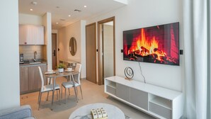 A modern living room with a TV displaying a fireplace, a dining area with a table and chairs, and a kitchen in the background. - Yogi - Bright Apartment With Panoramic CityScape Views (Dubai)
