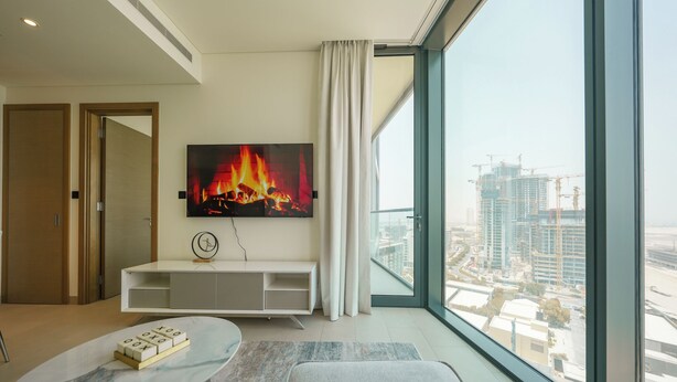 A modern living room with a large TV, a marble coffee table, and a view of a cityscape through large windows. - Yogi - Bright Apartment With Panoramic CityScape Views (Dubai)