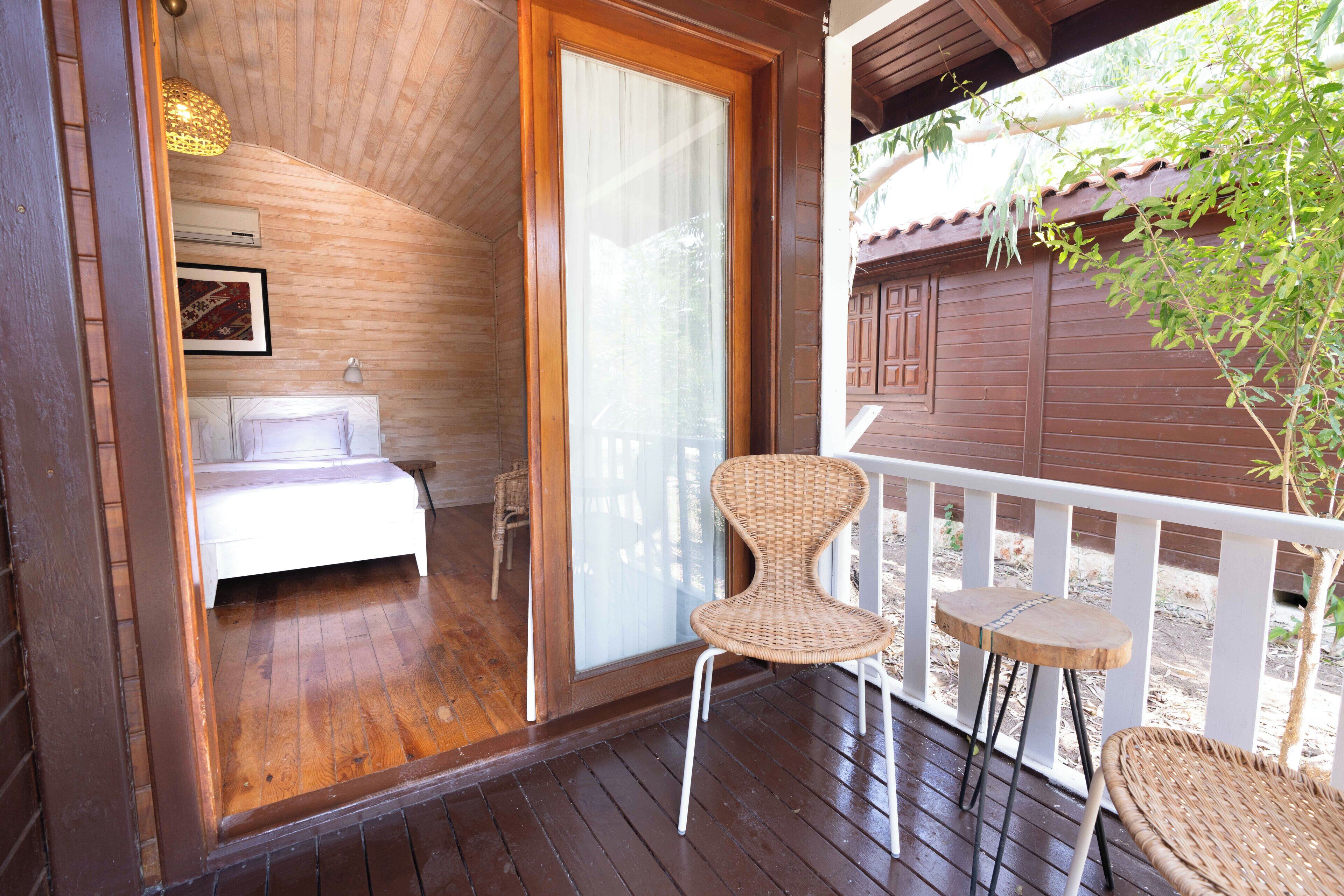 Un dormitorio con piso de madera, una cama, una silla blanca y una mesita en un balcón con vista a una cerca de madera y follaje.