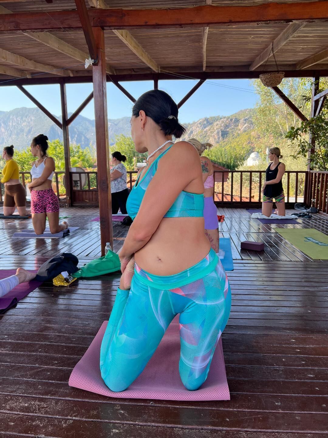 Un espacio al aire libre techado con suelo de madera, colchonetas de yoga y personas realizando actividades de yoga.