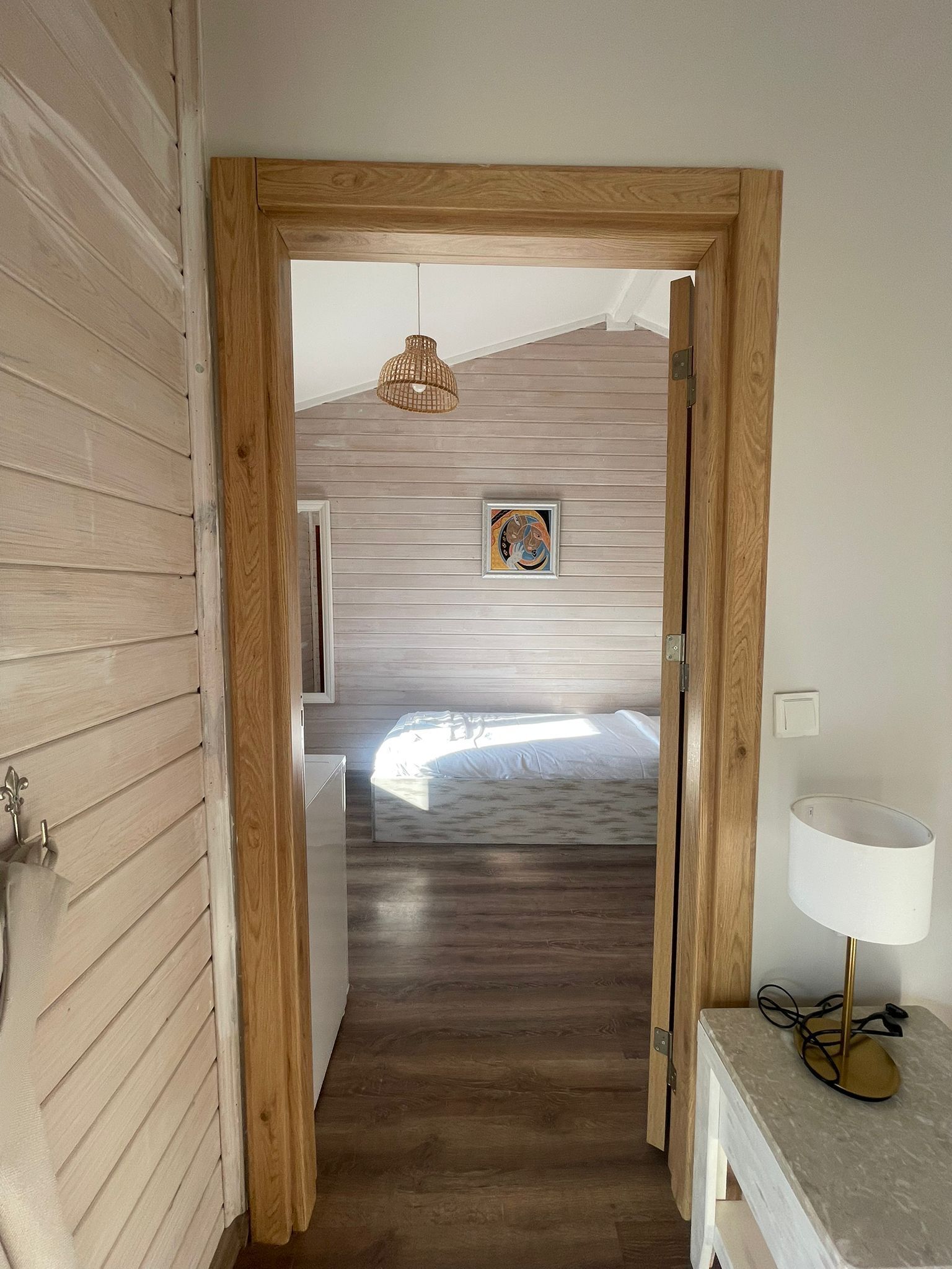 Un pasillo con paredes de madera, una cama con colchón blanco, una mesita de noche con lámpara y un aplique.