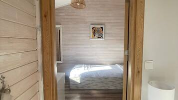 Un pasillo con paredes de madera, una cama con colchón blanco, una mesita de noche con lámpara y un aplique.