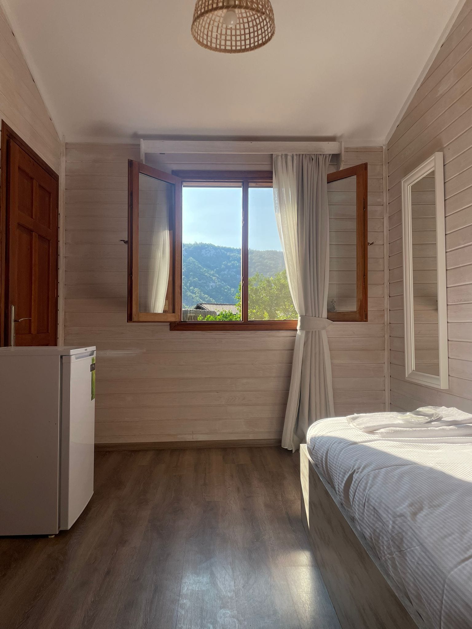 A bedroom with a wooden floor, a bed, a window with curtains, and a view of greenery outside.