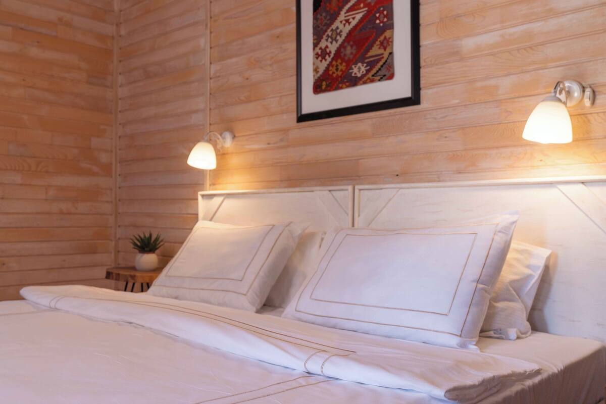 A neatly made bed with white linens and pillows, a small bedside table with a plant, and a framed artwork on the wall.