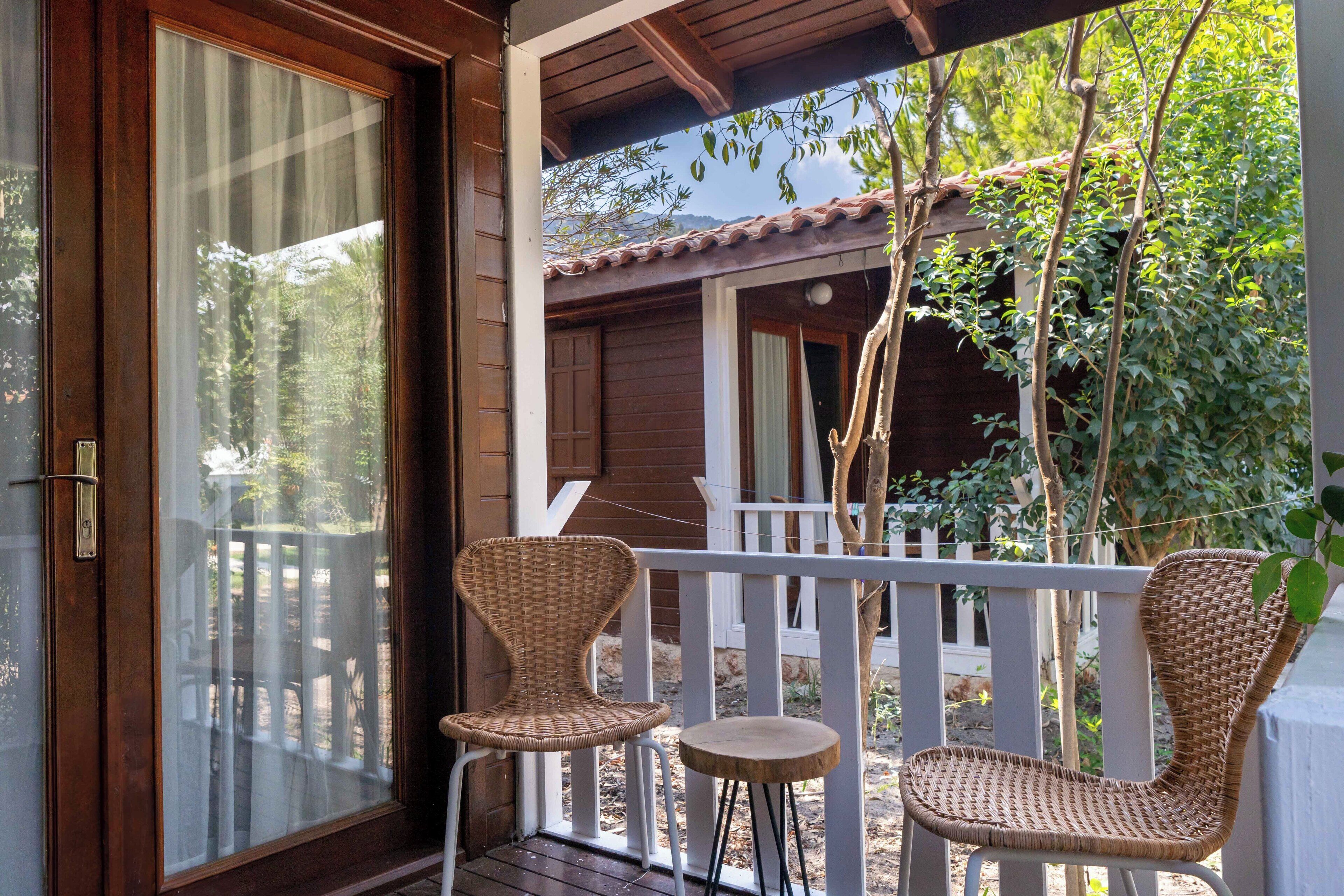Una terraza de madera con dos sillas de mimbre y una mesa pequeña, con vistas a un jardín con cerca blanca y árboles.