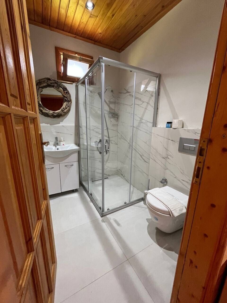Un baño con cabina de ducha de vidrio, inodoro, lavabo y techo de madera.