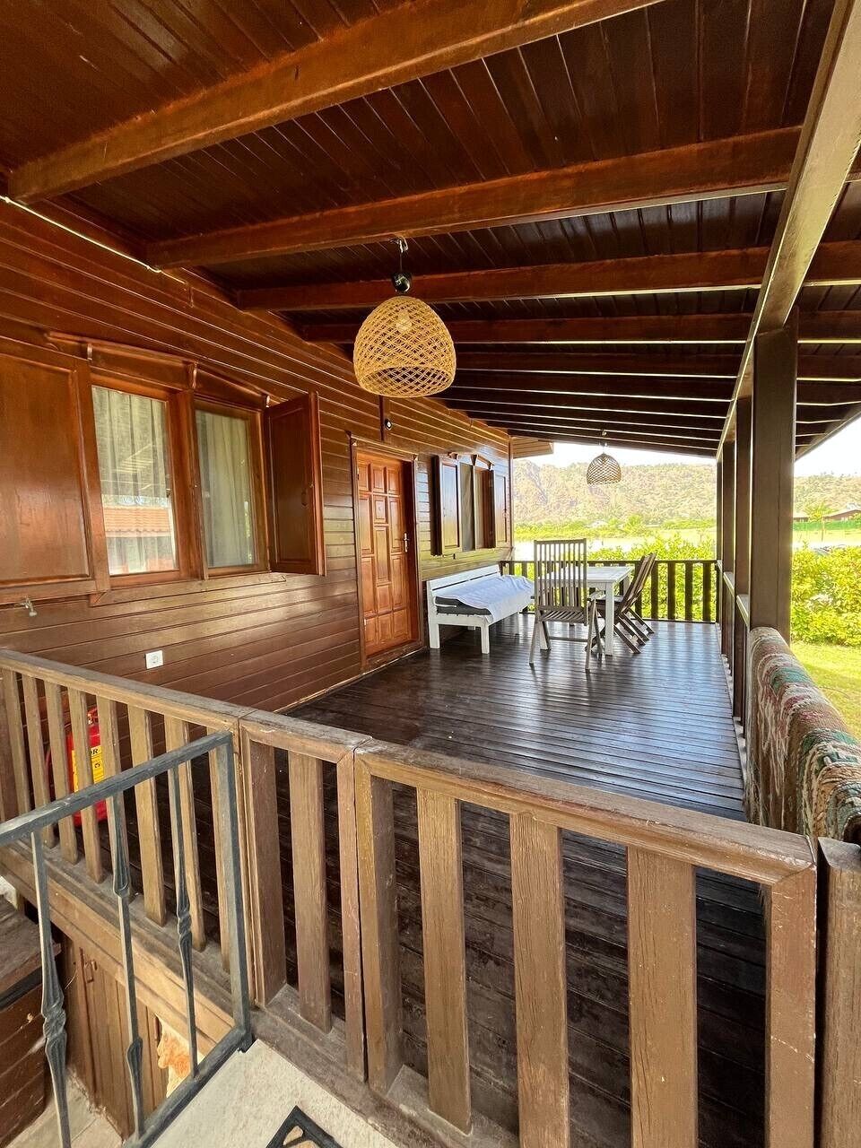 A wooden deck with a table and chairs, a hanging light fixture, and a view of greenery.