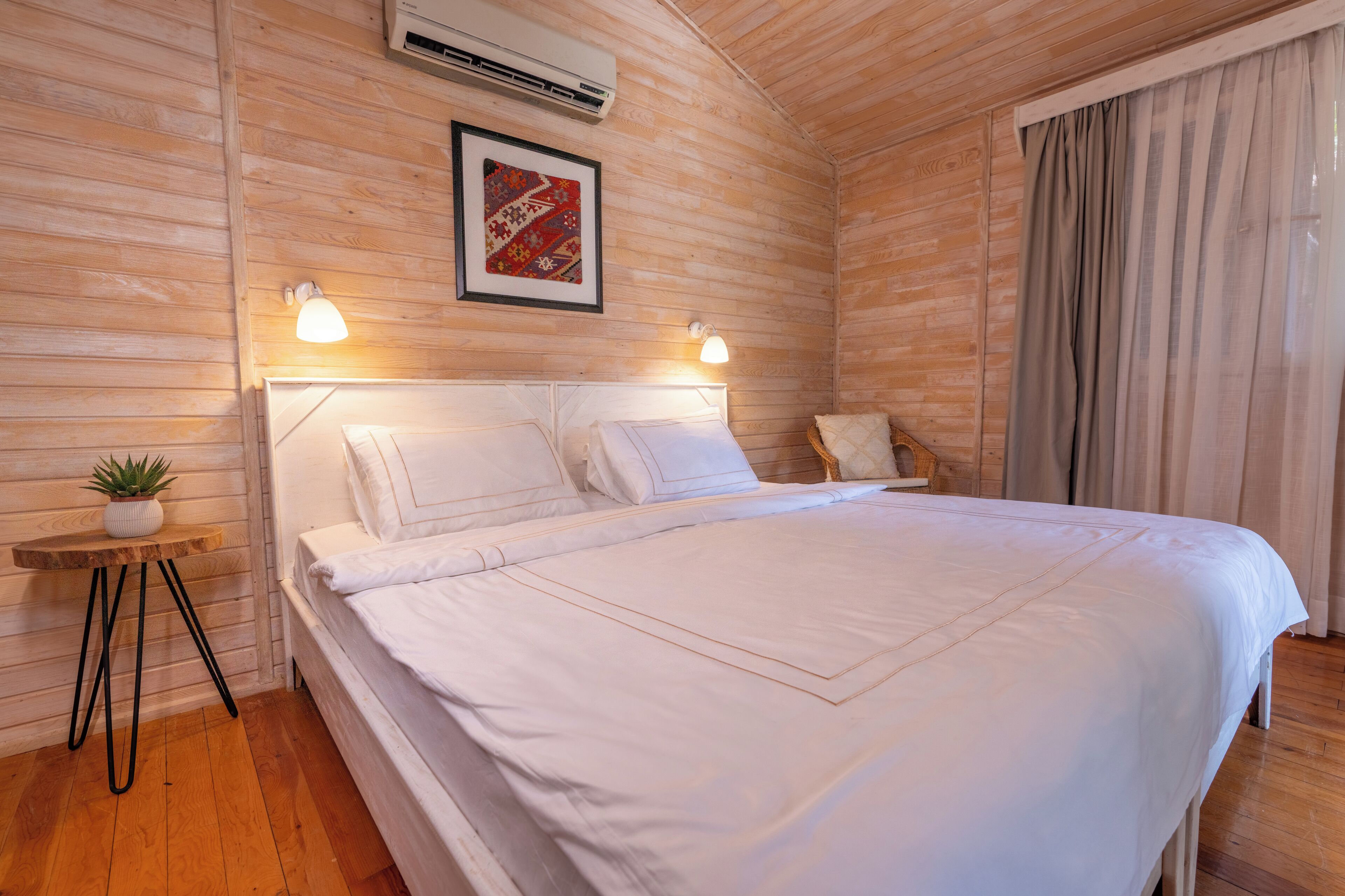A neatly made bed with white linens, a small bedside table with a plant, and a framed picture on the wall.