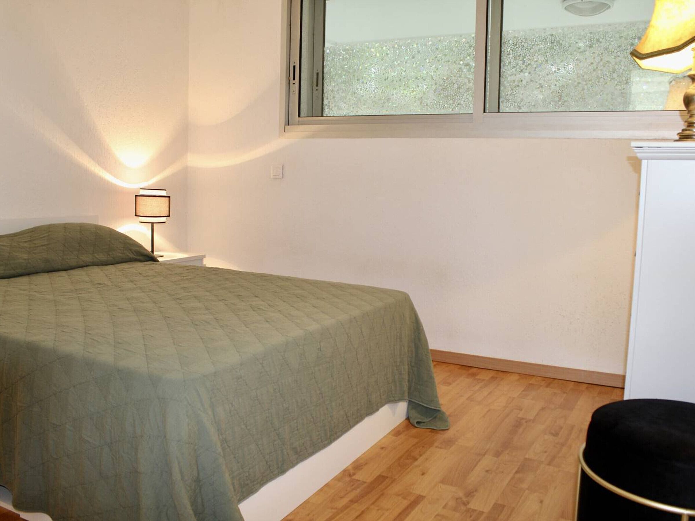 A small, neatly arranged bedroom with a bed, a nightstand, a lamp, and a window.