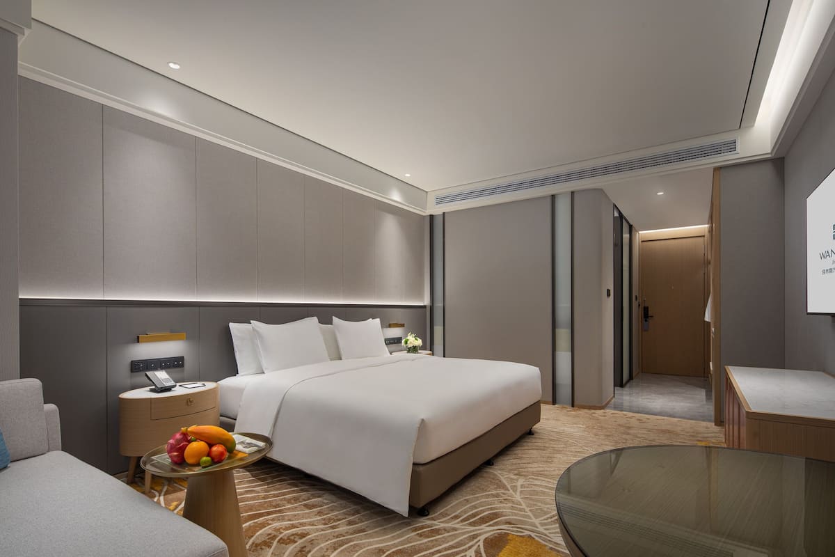 A modern hotel room with a large bed, a bedside table with a fruit bowl, a small desk, and a glass coffee table.