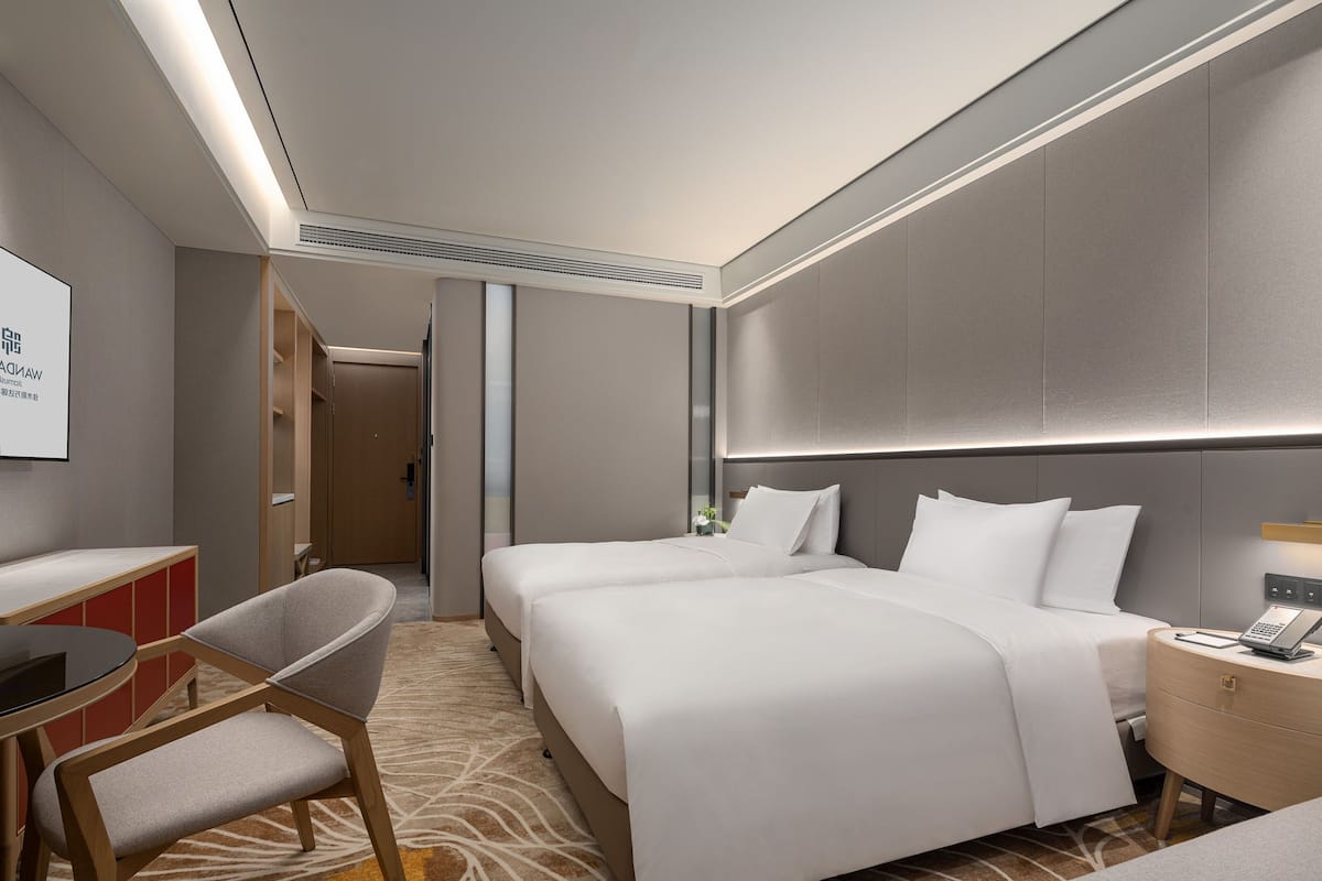 A hotel room with two beds, a desk, a chair, and a bedside table with a lamp.