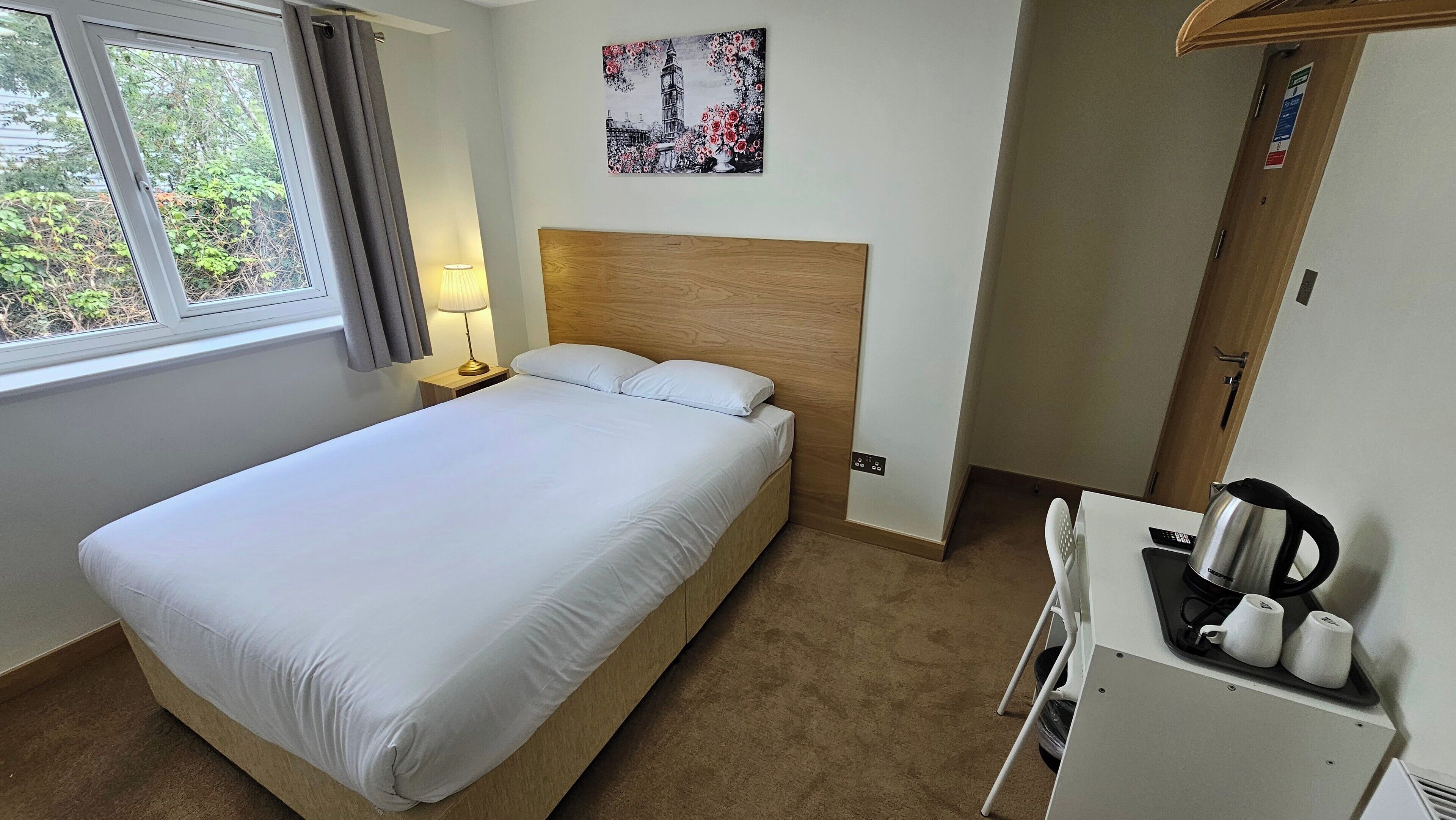 A hotel room with a single bed, a nightstand with a lamp, a painting of a famous landmark, a wooden headboard, a small kitchenette with a kettle, and a window with curtains.
