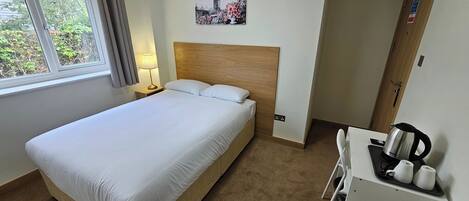 A hotel room with a single bed, a nightstand with a lamp, a painting of a famous landmark, a wooden headboard, a small kitchenette with a kettle, and a window with curtains.
