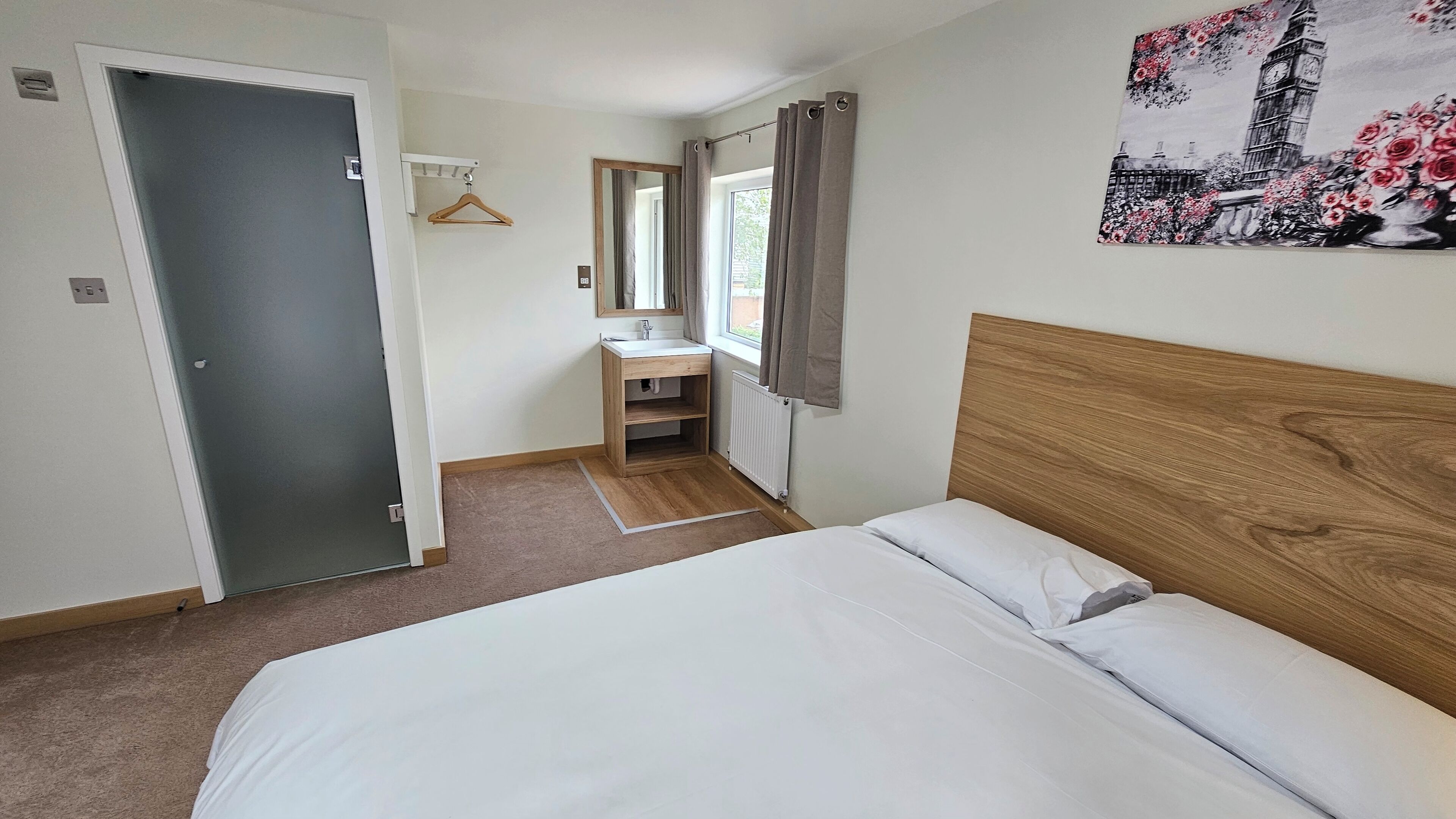 A bedroom with a wooden headboard, a bed, a bedside table, a mirror, a window with curtains, and a bathroom with a sink and mirror.