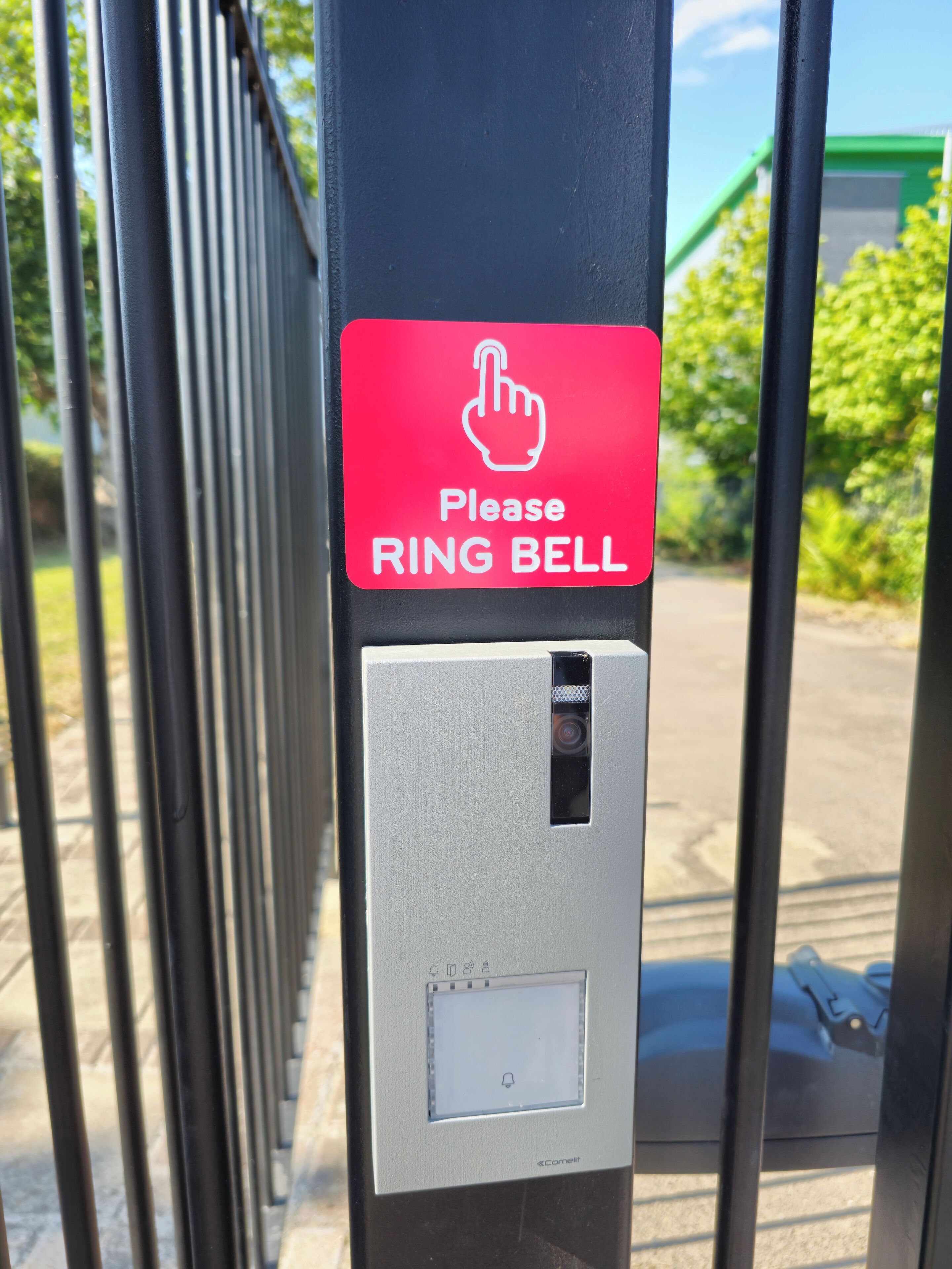 A security gate with a red sign instructing to ring the bell, a keypad, and a camera.