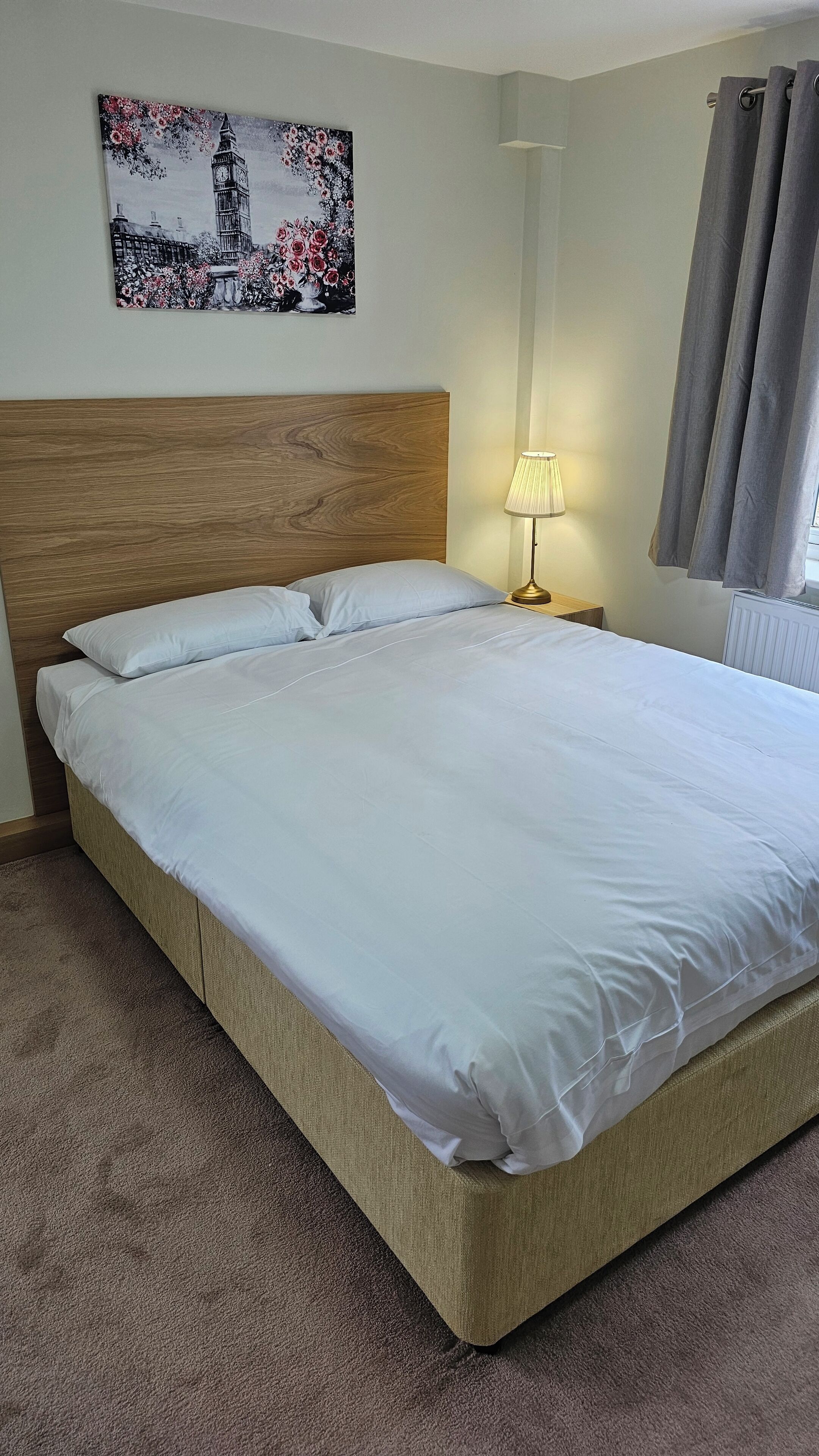 A neatly made bed with white linens in a room with a wooden headboard, a wall-mounted lamp, and a painting of a cityscape.
