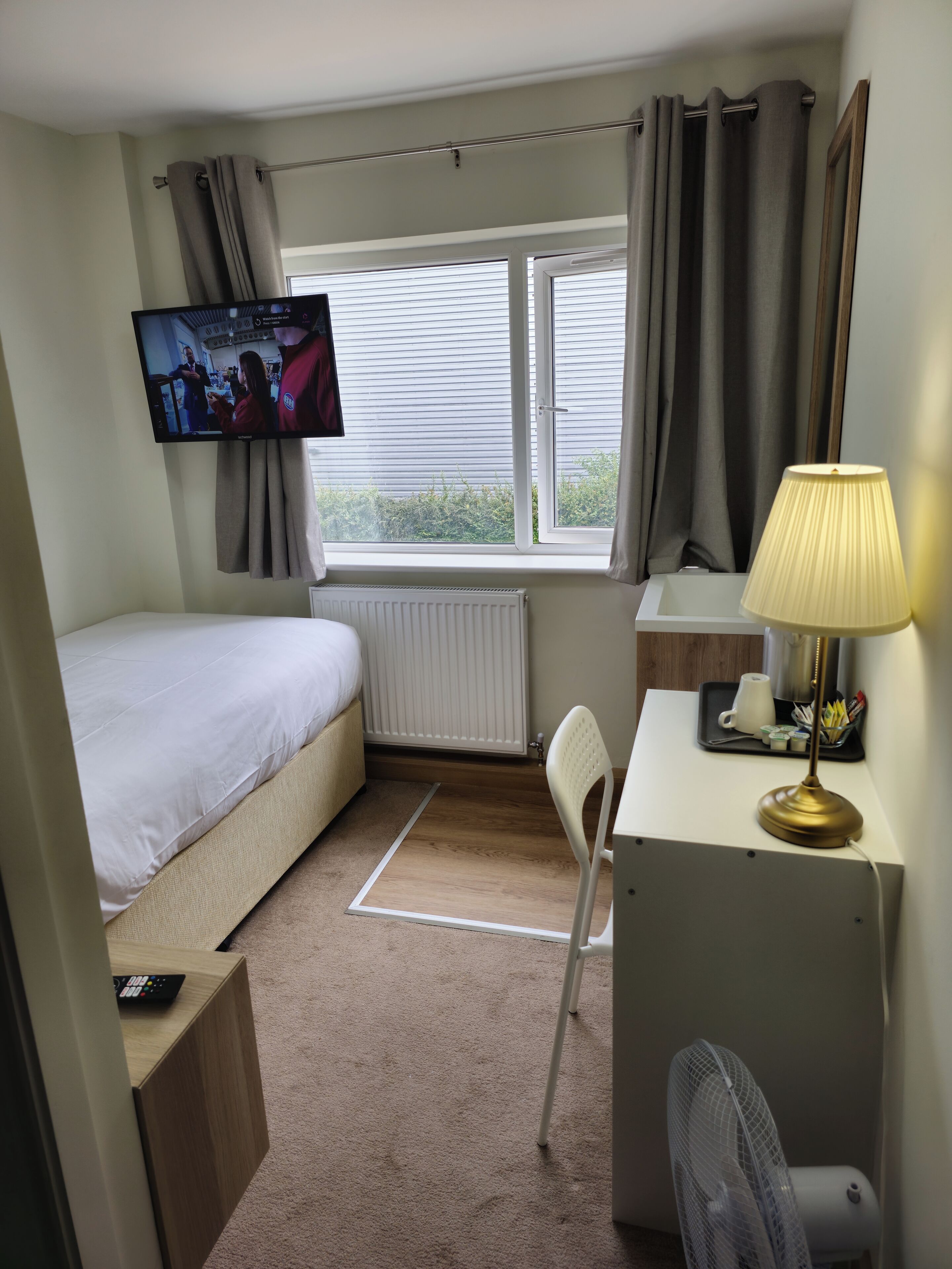 A compact hotel room with a single bed, a desk with a lamp, a chair, a TV mounted on the wall, and a window with curtains.
