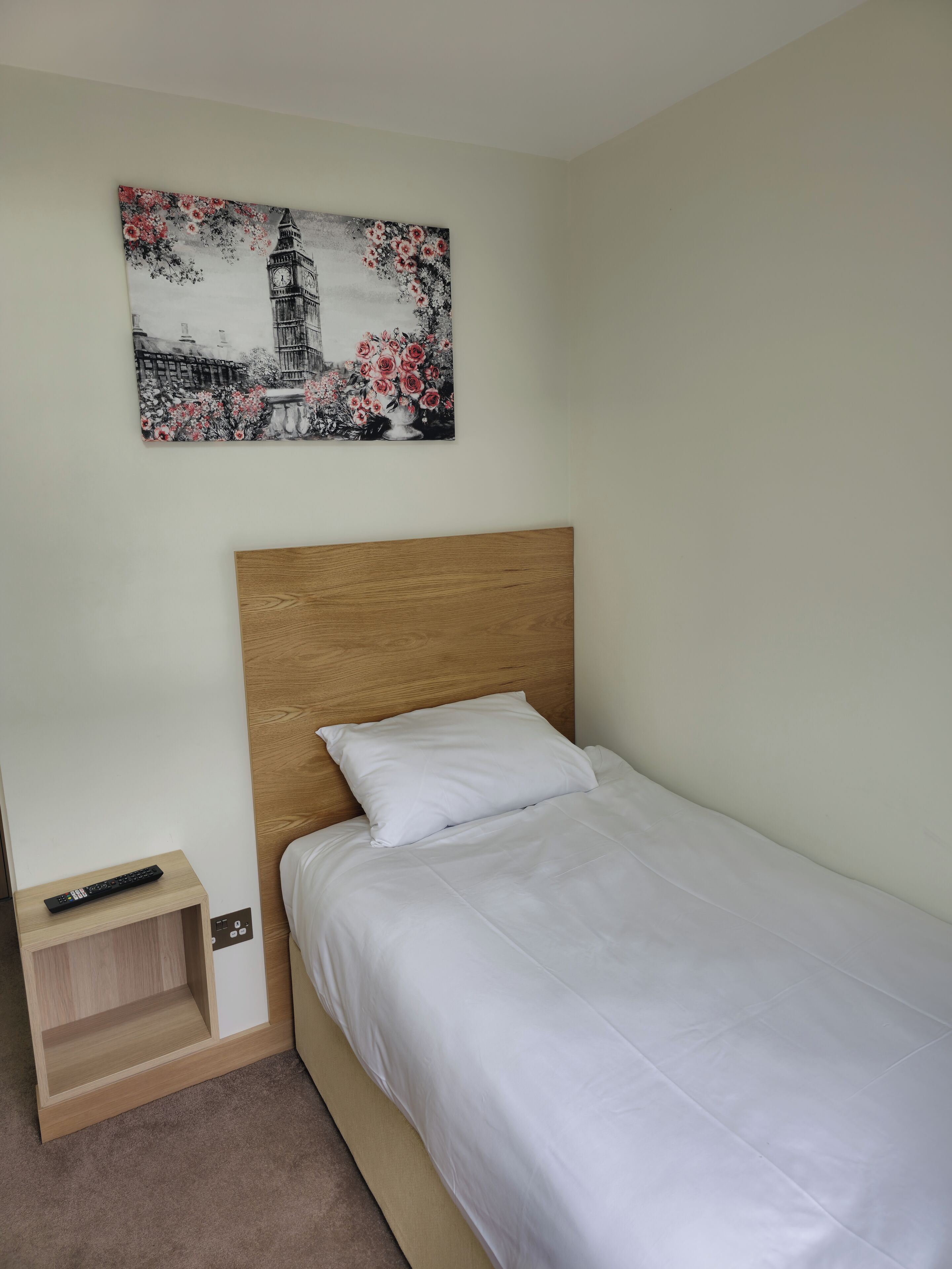 Ein Einzelbett mit Holz-Kopfteil, eine Fernbedienung auf einem Nachttisch und ein Bild einer Turuhr und Blumen an der Wand.