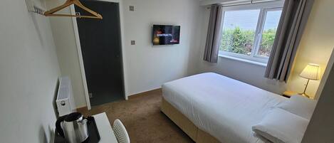 A hotel room with a bed, a TV, a desk with a kettle, a chair, and a window with curtains.