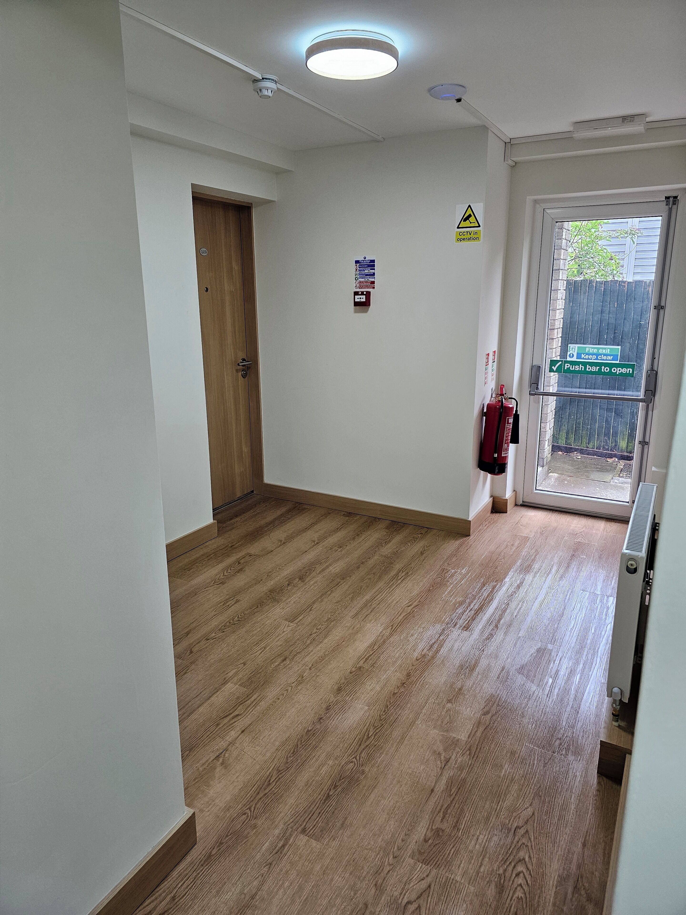 Un couloir avec un plancher en bois, une porte, un panneau indiquant la sortie de secours et un panneau indiquant la présence d’un système de vidéosurveillance.