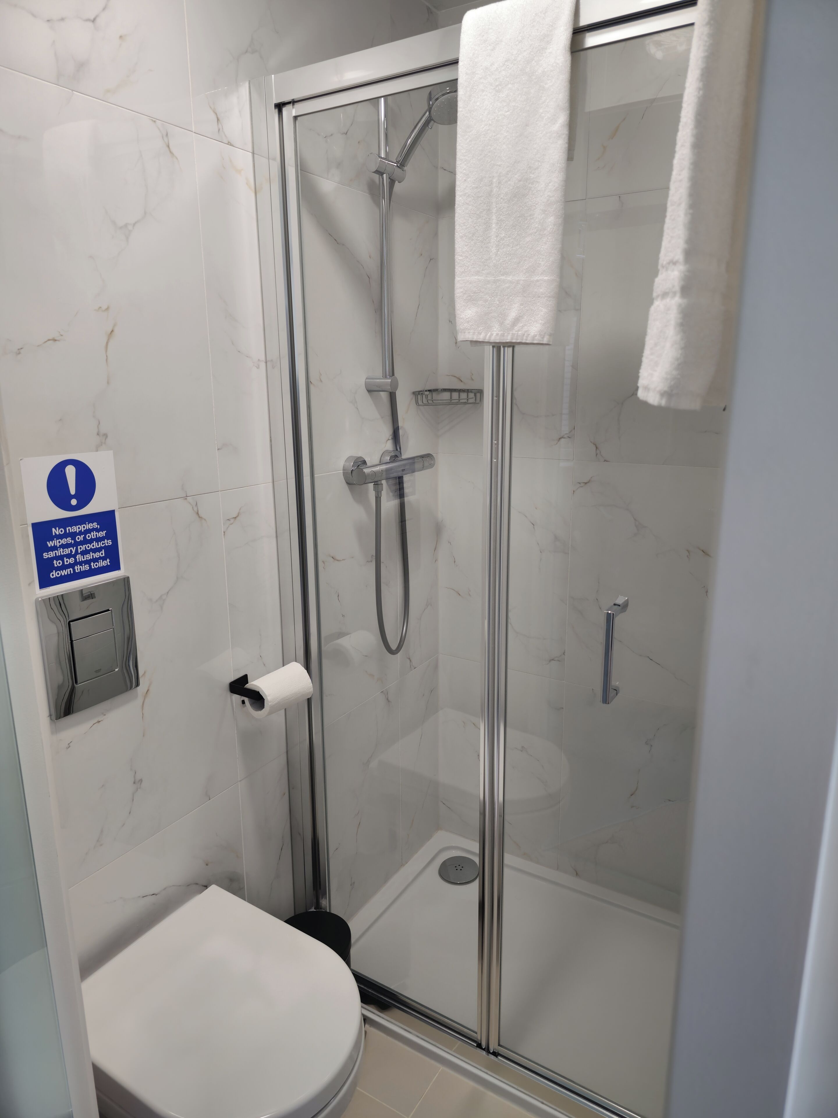 A glass-enclosed shower stall with a handheld showerhead, a towel hanging on the door, and a sign prohibiting the disposal of nappies, wipes, or other sanitary products down the toilet.