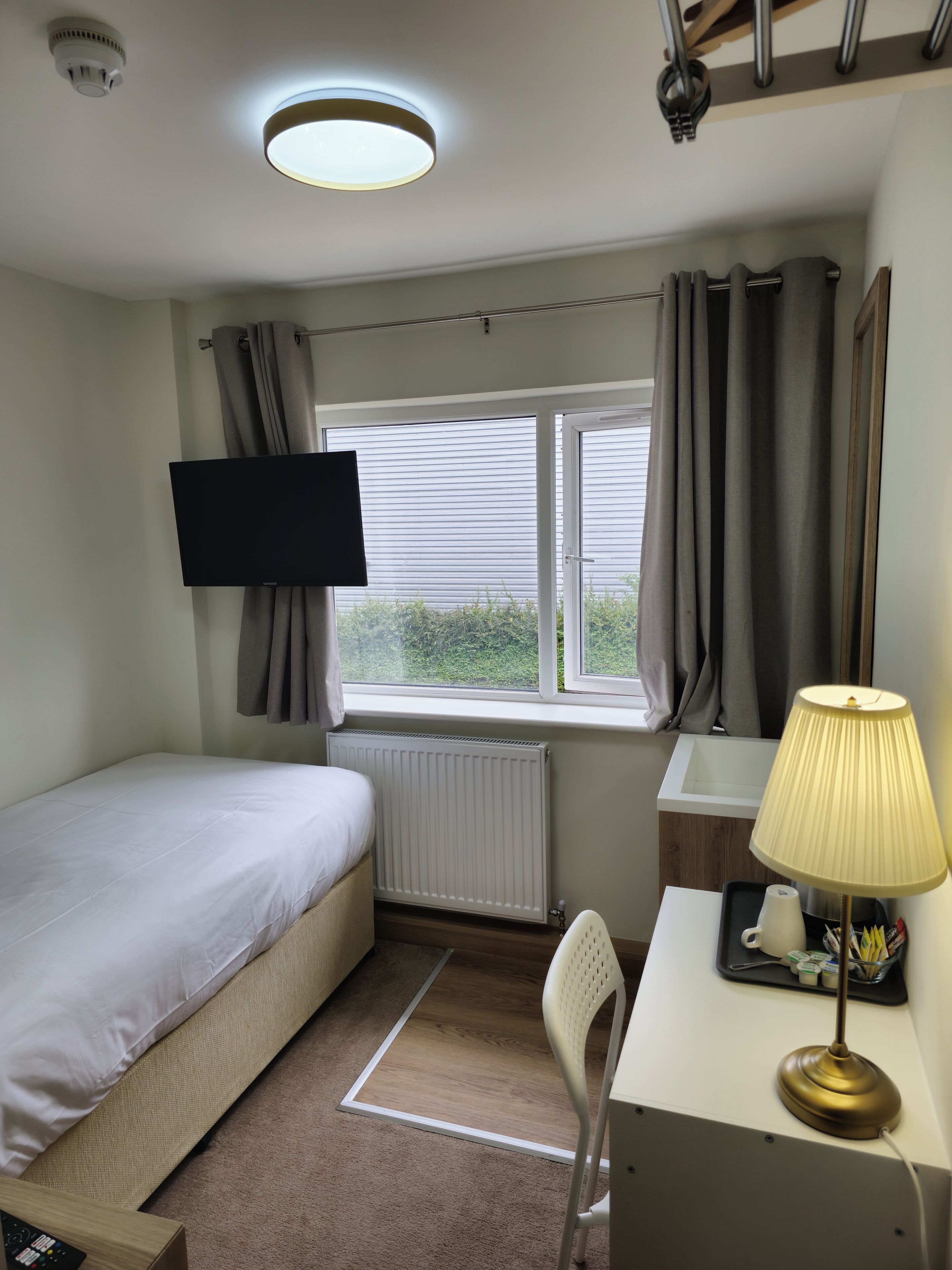 Un dormitorio con cama, escritorio con lámpara, televisor, radiador y ventana con cortinas.