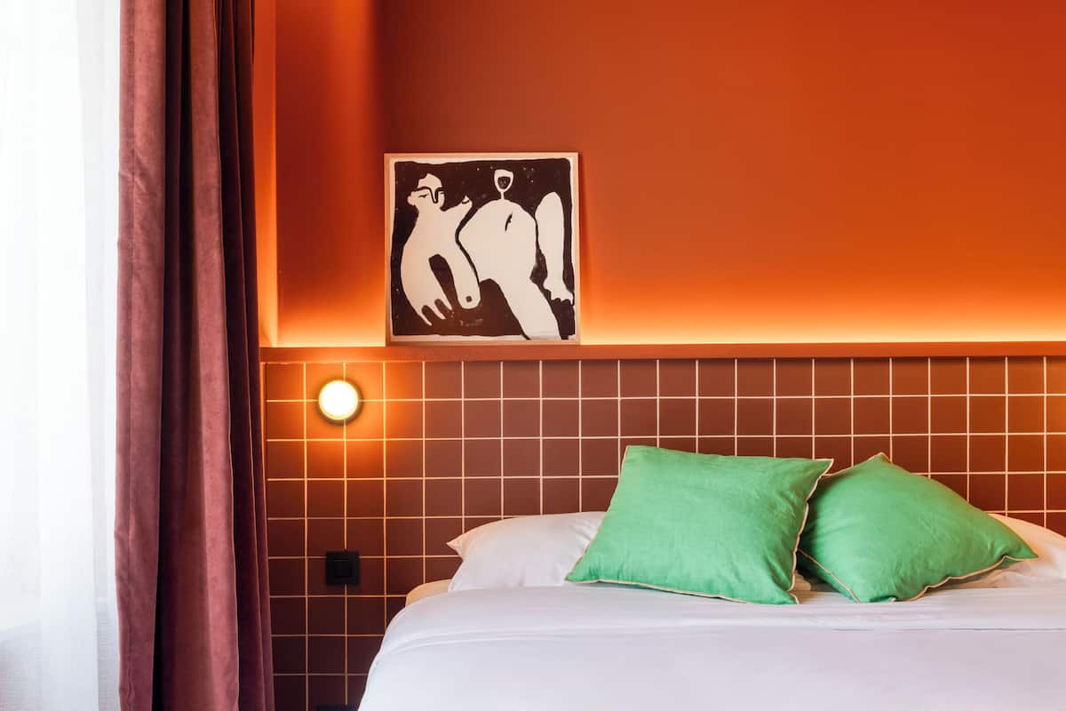 A hotel room with a bed, green pillows, a bedside table with a book and a candle, and a wall with a geometric light fixture.