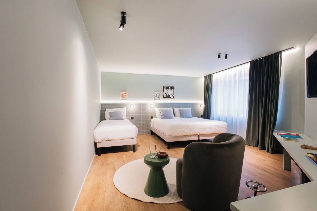A modern hotel room with two beds, a desk, a chair, and a small side table.