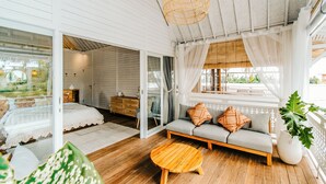 Um quarto espaçoso com piso de madeira, um sofá com almofadas, uma mesa de centro redonda de madeira e uma grande janela com cortinas leves.