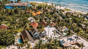 Vista aérea de uma estância costeira com vários edifícios, palmeiras e uma praia.