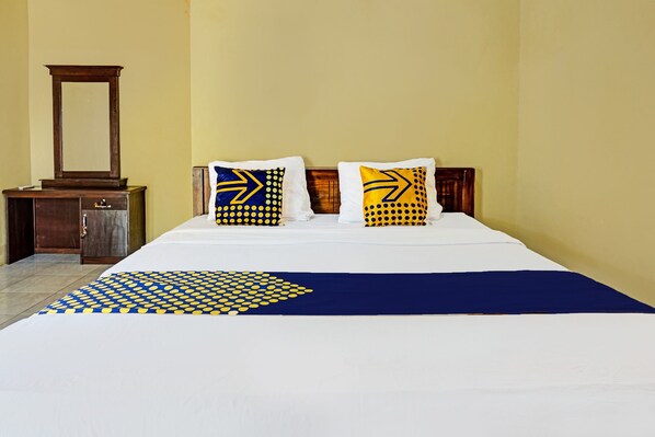 Una cama bien hecha con una colcha azul y amarilla estampada, dos almohadas con diseños geométricos y una mesita de noche de madera con espejo.