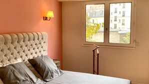 A bedroom with a tufted headboard, a bed with grey bedding, a standing lamp, and a window with a view of buildings and greenery. - Centre Paris, Grand Appartement Calme et Lumineux (Paris)
