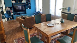 A modern living room with a wooden dining table, green upholstered chairs, a blue sofa, and a bookshelf filled with books and decorative items. - Centre Paris, Grand Appartement Calme et Lumineux (Paris)
