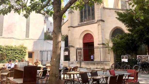 An outdoor café with tables and chairs, a tree, and a building with a red door in the background. - Centre Paris, Grand Appartement Calme et Lumineux (Paris)