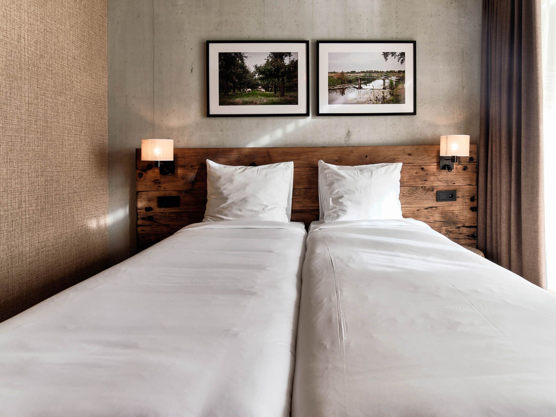 A double bed with white linens, a wooden headboard, two wall-mounted lamps, and two framed pictures on the wall.