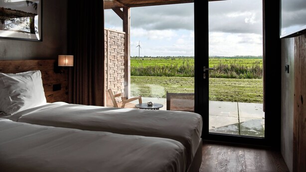 Un dormitorio con una cama grande, cabecera de madera, mesita de noche, lámpara y vista a un campo verde con una turbina eólica.