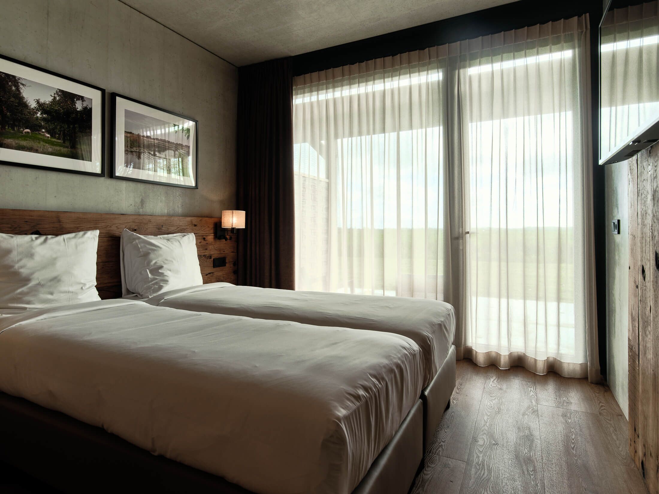 Een hotelkamer met twee bedden, een houten hoofdbord, een nachtkastje met een lamp en grote ramen voorzien van dunne gordijnen.