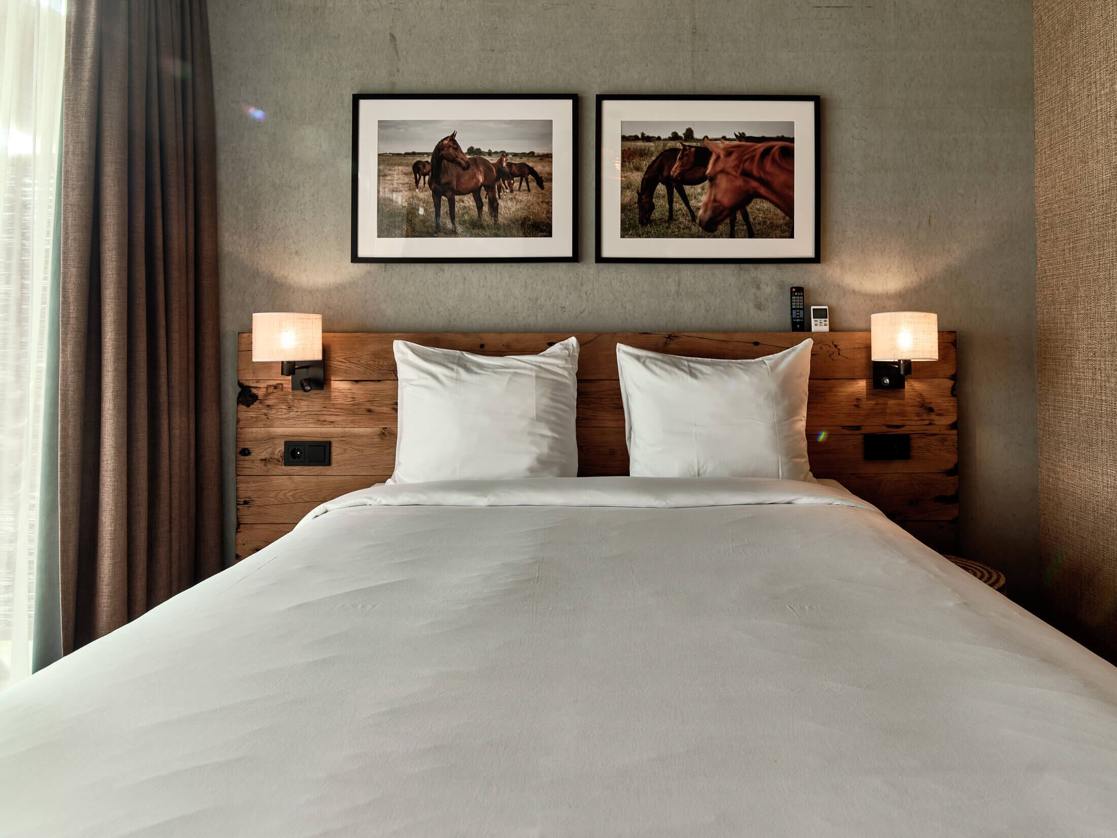 Habitación de hotel con cabecera de madera, dos cuadros enmarcados de caballos y lámparas de noche.