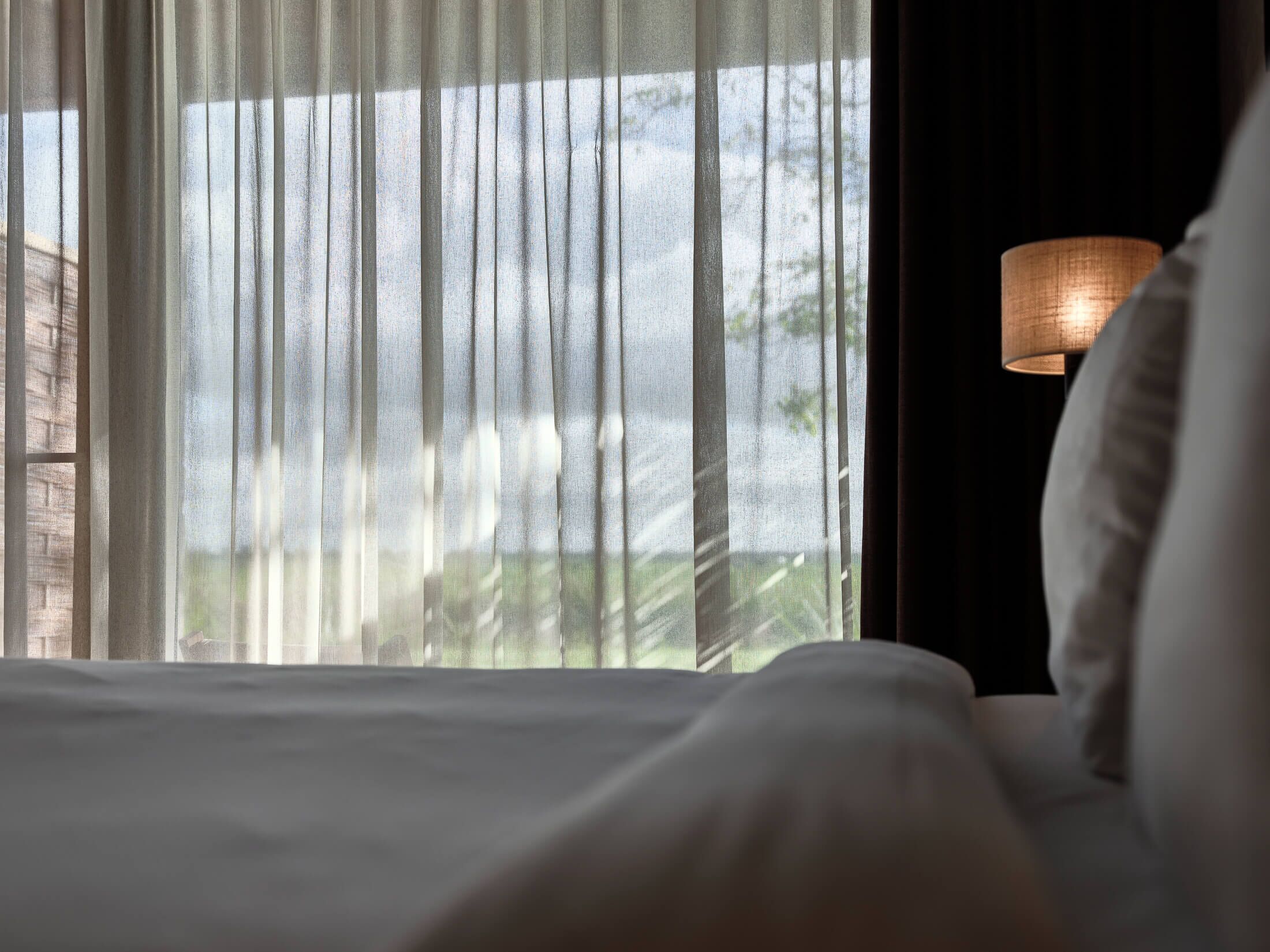 A hotel room with a bed, sheer curtains, and a lamp.